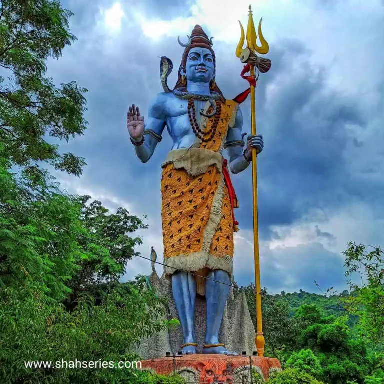 The image is a statue of a Shiva holding a staff. It is an outdoor sculpture located near a building or temple. The Shiva statue is surrounded by trees and plants, with the sky and clouds in the background.