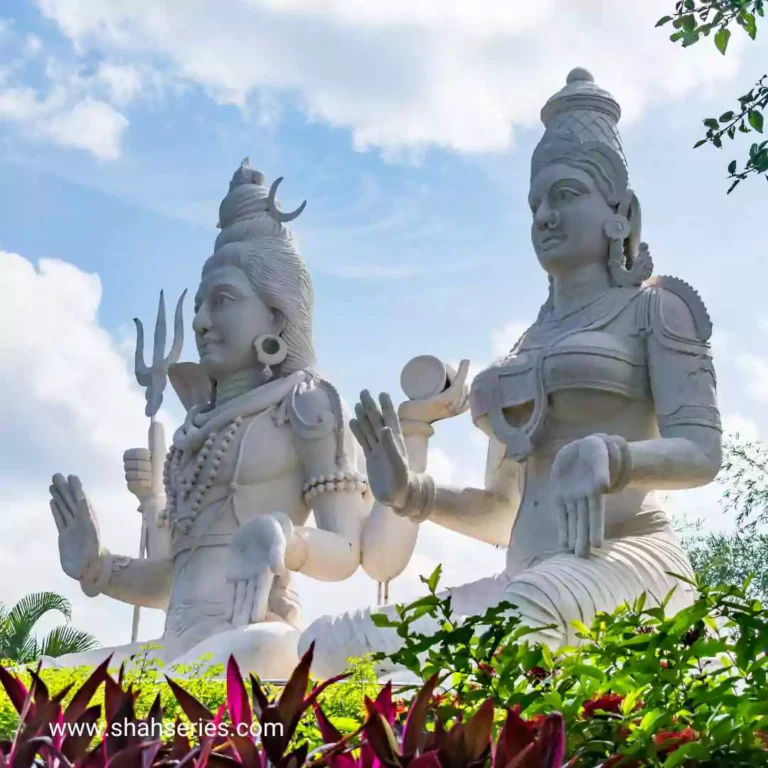 This is a photo of Shiv Parvati holding a sword. It is located outdoors, possibly in a temple or a place of worship. The background includes a clear blue sky with clouds, and there are trees and other plants surrounding the area.
